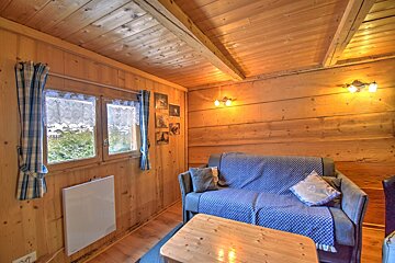 Rustic wooden cabin interior with a blue sofa, coffee table, and two windows looking out. Warm lighting adds to the cozy feel.