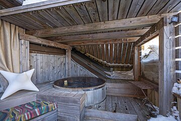 A rustic wooden outdoor area features a hot tub, bench with a star pillow, and icicles. Snow is visible outside, suggesting a cozy winter setting.