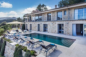 A luxurious stone villa with a sparkling pool and multiple lounge chairs, set against a backdrop of rolling green hills under a bright blue sky.