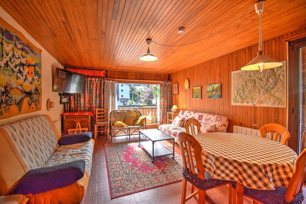 A cozy, wood-paneled living room with a dining area, sofa, coffee table, and balcony access. Features a tapestry, paintings, and a checkered tablecloth.