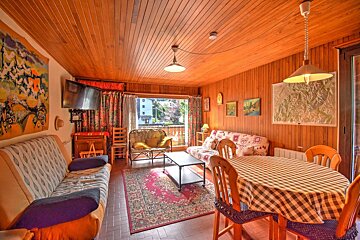 A cozy, wood-paneled living/dining room with rustic sofas, a checkered-clothed dining table, various artworks, and a balcony view.