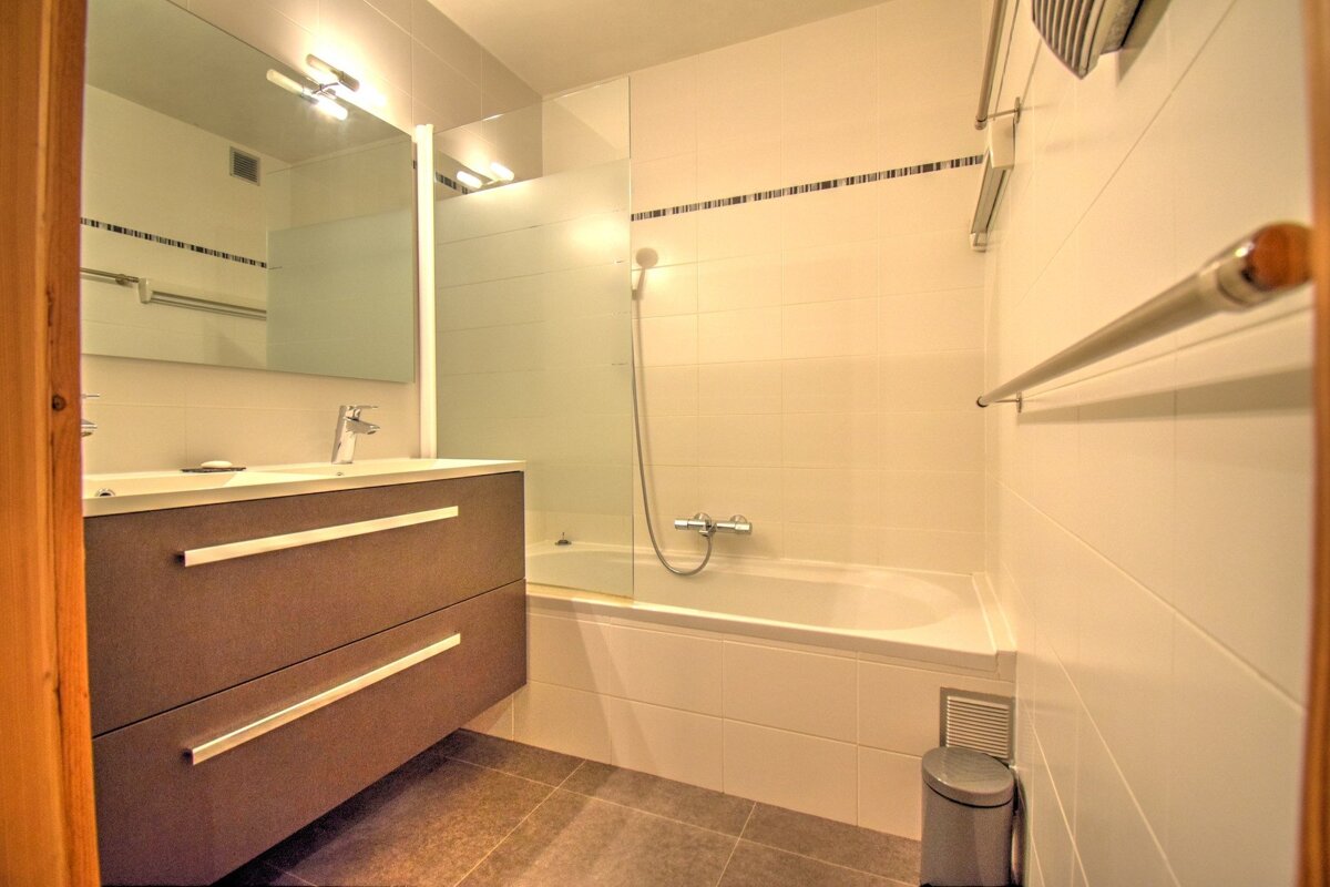 A modern bathroom featuring a brown floating vanity with sink, mirror, and a bathtub with shower. White tiled walls, dark floor.
