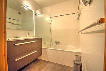 A modern bathroom featuring a brown floating vanity with sink, mirror, and a bathtub with shower. White tiled walls, dark floor.
