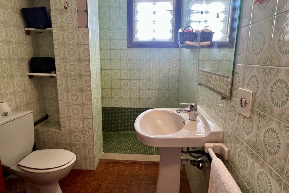 A green-tiled bathroom with a toilet, pedestal sink, and shower area. It features light green patterned wall tiles, dark green mosaic in the shower, and a window.