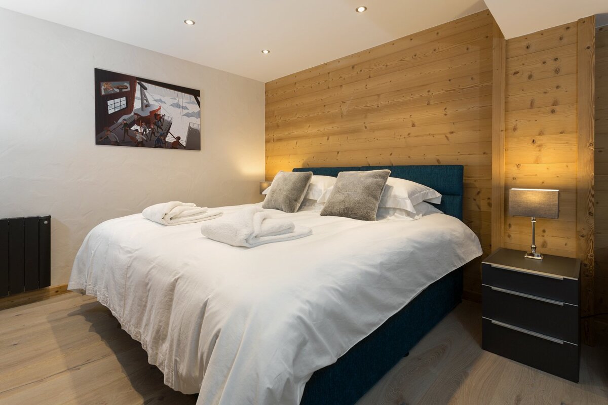 A modern, cozy bedroom with a large bed, crisp white linen, and a wooden feature wall. An abstract painting hangs above, and a lamp lights the nightstand.