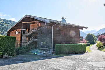 Large wooden chalet with external stairs, surrounded by green hedges, under a sunny sky with mountains in the background.
