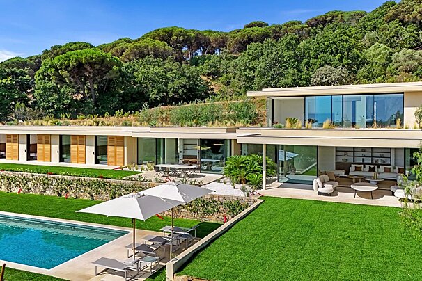 Luxurious modern house with a pool, expansive lawn, outdoor living areas, and a verdant hillside backdrop under a blue sky.