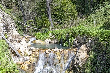 Cascading waterfalls flow into clear rock pools in a lush green natural setting, framed by stone walls and a distant forest.