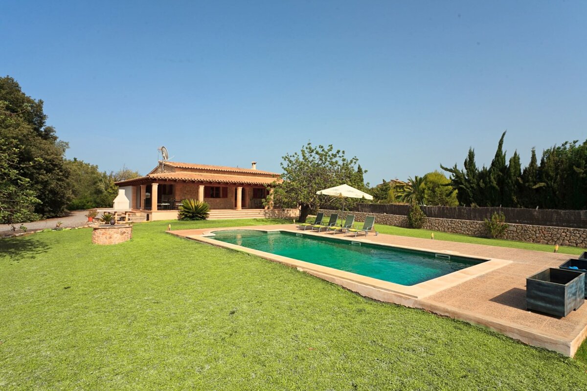 A sunny villa with a tiled roof, surrounded by a lush green lawn and a refreshing blue swimming pool with lounge chairs under a clear sky.