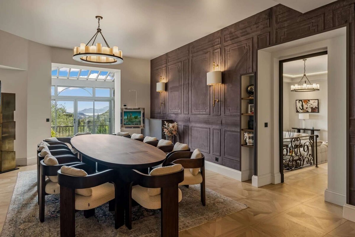 An elegant dining room with a large oval table, modern chairs, a dark paneled wall, and a stunning mountain and water view through a panoramic window.