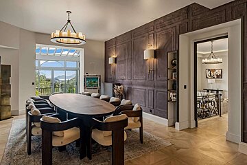 An elegant dining room features a large oval table with numerous chairs, a round chandelier, and a scenic view through a massive window.