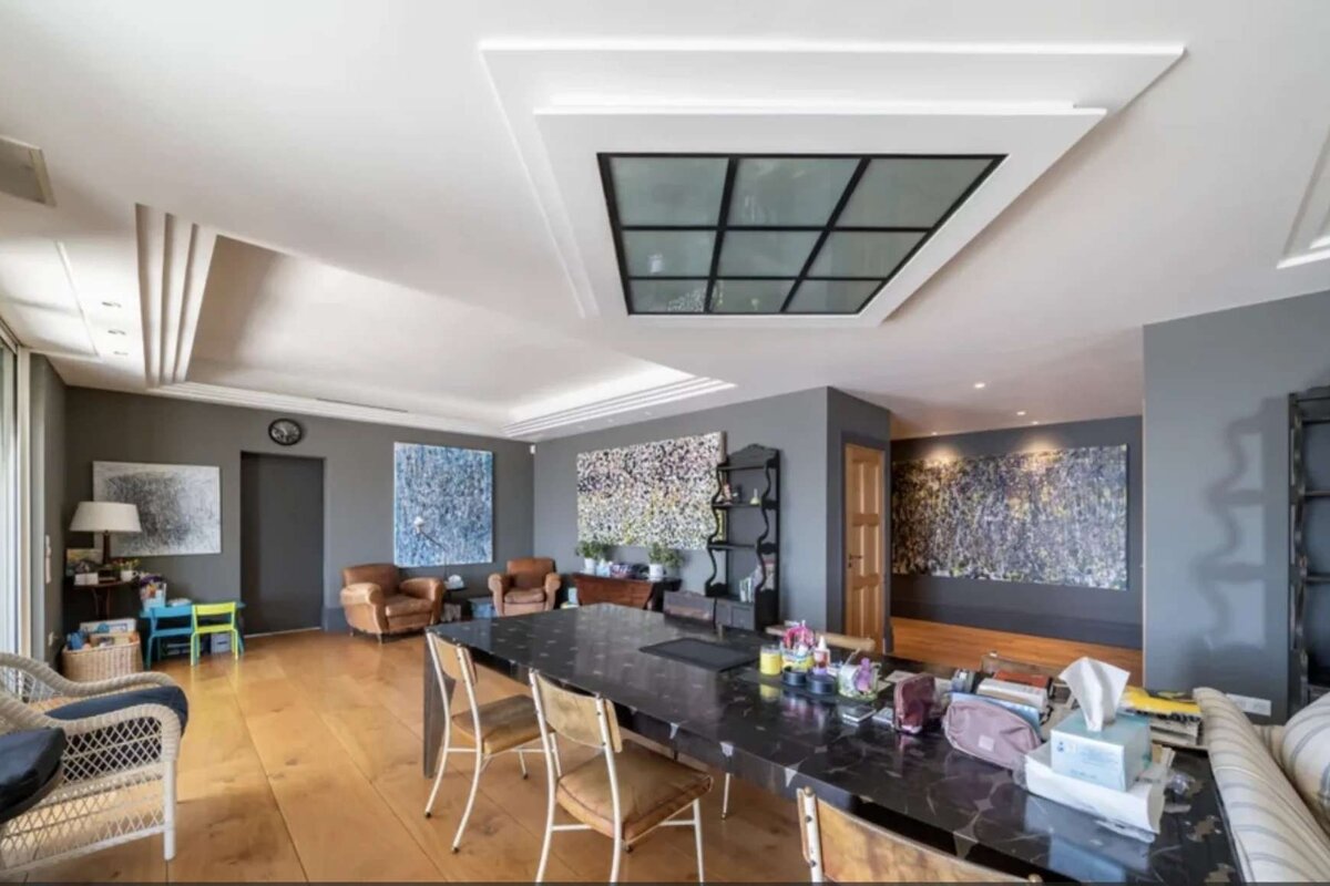 A spacious, modern room with dark gray walls, wooden floor, and a prominent ceiling light panel. Features abstract art, a long dining table, and lounge seating.