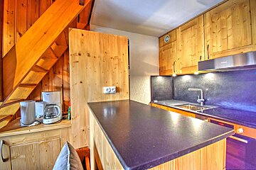 A cozy, wooden kitchen with a dark counter, sink, stove, and range hood. Coffee maker and kettle sit next to a wooden staircase.