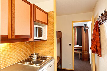 A compact studio apartment featuring a kitchenette with orange cabinets, microwave, and stove, next to a bunk bed and a doorway to another room.