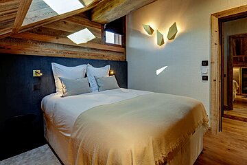 A rustic-modern bedroom features a wooden beamed ceiling, sleek geometric wall lights, and a comfy white bed with a beige throw.
