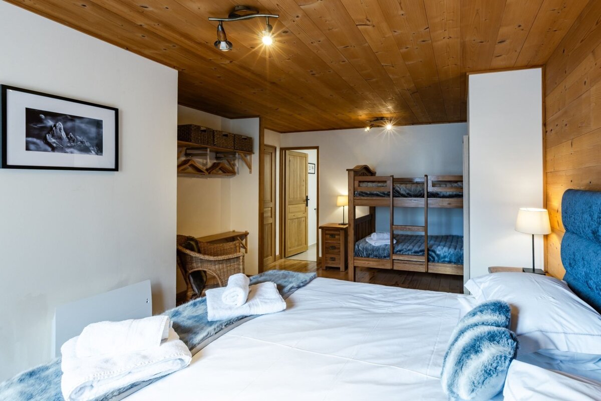 Cozy rustic bedroom featuring a large bed with blue pillows, a wooden ceiling, and a bunk bed in the background.