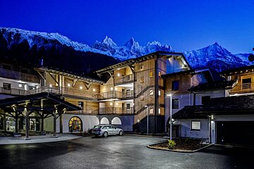 An illuminated wooden alpine hotel at night, with a parked car and a backdrop of snowy mountain peaks under a deep blue sky.