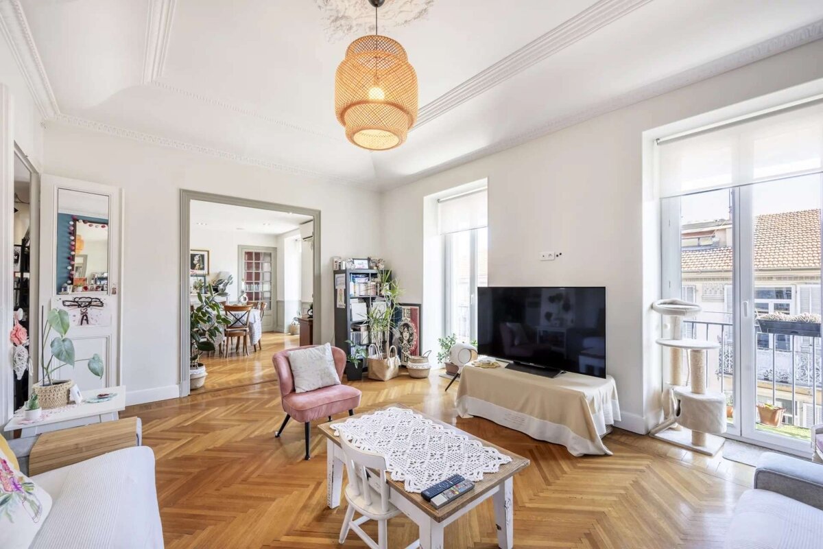 Spacious, bright living room featuring herringbone wood floors, white walls, a woven pendant lamp, TV, pink armchair, and a cat tree by the balcony door.