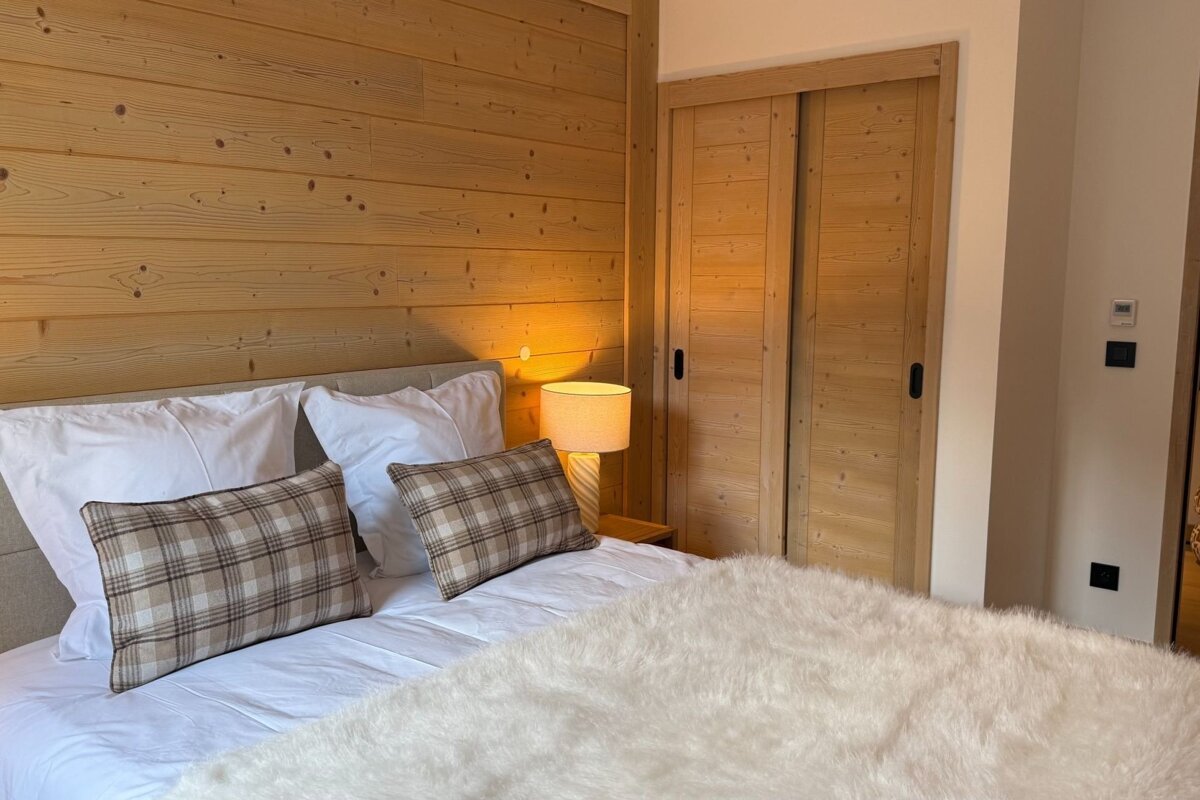 Cozy bedroom with light wood paneled walls, a bed featuring plaid pillows and a fluffy white blanket, and a glowing bedside lamp.