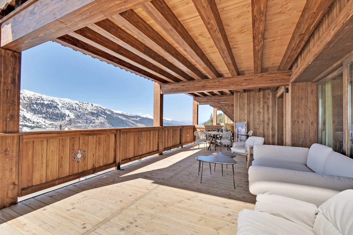 Spacious wooden balcony with white outdoor seating and stunning views of snow-capped mountains under a clear blue sky.