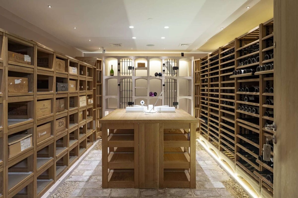 A modern wine cellar featuring extensive wooden racks filled with wine bottles and boxes, a central display table, and stone floors.