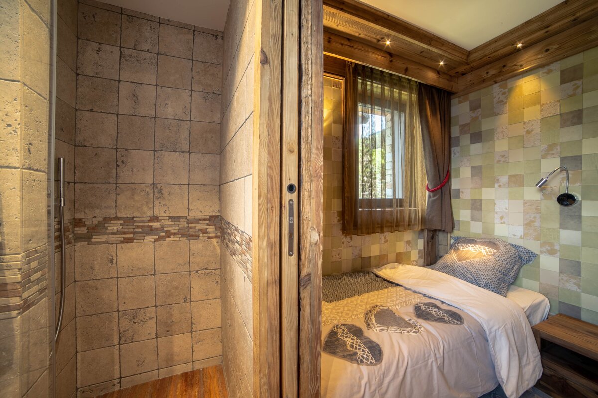 A cozy bedroom with a single bed and window, next to a tiled shower, separated by a rustic wooden partition. Warm lighting and wood accents.
