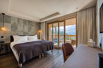 Modern chalet bedroom featuring a large bed, stone accent wall, and sliding doors leading to a balcony with stunning mountain views.