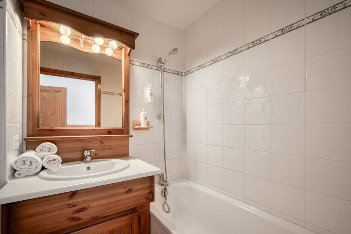 Cozy bathroom with a wooden vanity, matching framed mirror with lights, and a bathtub set against white tiled walls.