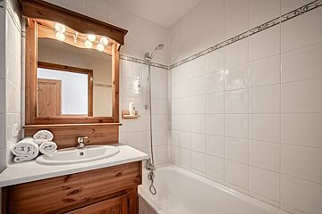 Cozy bathroom with a wooden vanity, matching framed mirror with lights, and a bathtub set against white tiled walls.