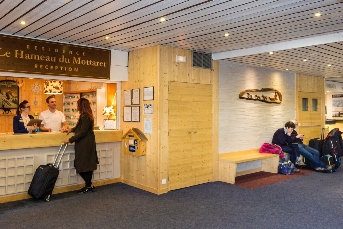 A busy hotel reception: staff assist a guest with luggage, while two others wait on a bench with bags in a cozy, wood-paneled lobby.