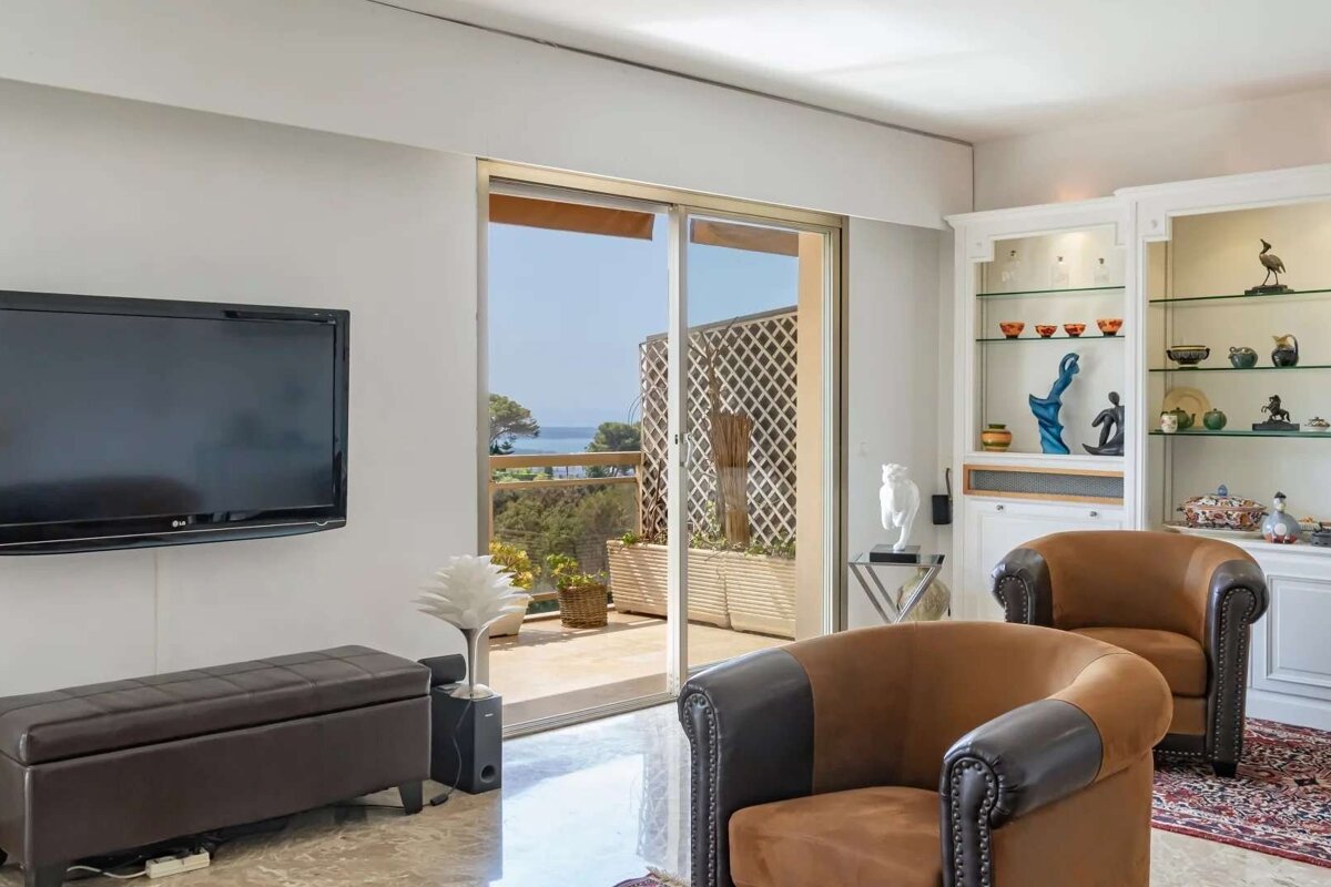 A bright living room with a TV, brown armchairs, and a balcony offering a clear view of the sea. Decorative cabinets line a wall.