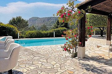 Sunny pool area with a stone patio, vibrant pink flowers on a pergola, and majestic mountains under a clear blue sky.