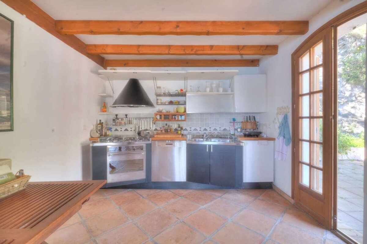 A modern kitchen features exposed wooden beams, terracotta floor tiles, stainless steel appliances, and a black range hood, with a French door opening outside.