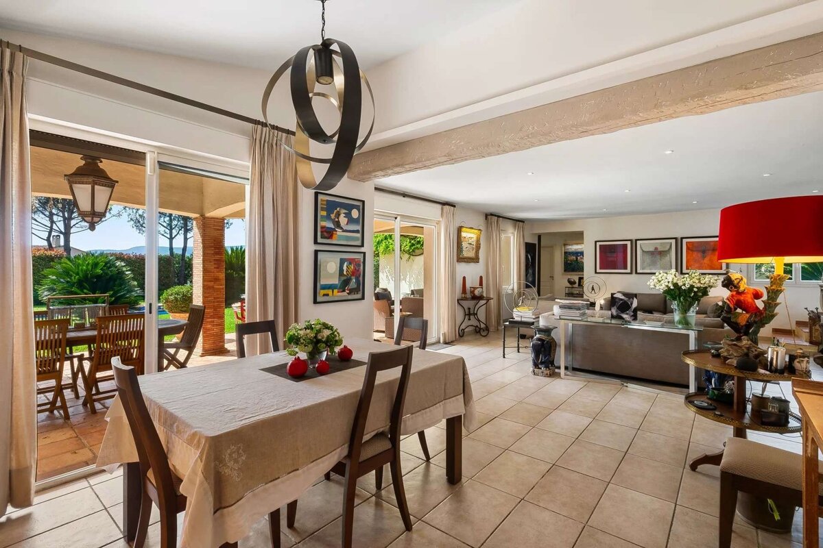 A bright, open-plan living and dining room with tiled floor, overlooking a garden patio. Features a dining table, chandelier, sofa, and art.