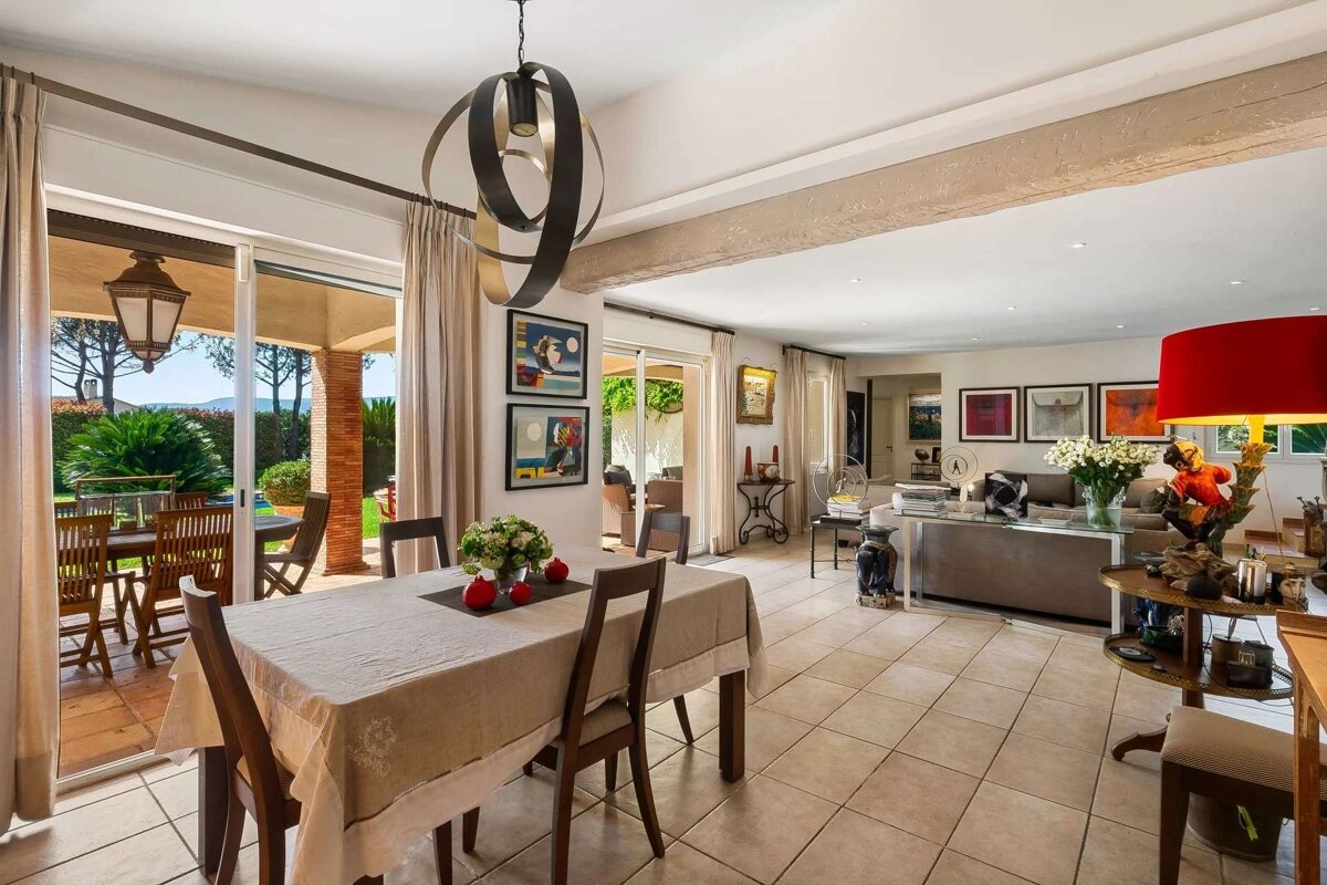 A bright, open-plan dining and living room with a large glass door leading to a sunny outdoor patio and lush garden. Contemporary decor.