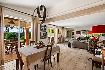 A bright, open-plan dining and living room with a large glass door leading to a sunny outdoor patio and lush garden. Contemporary decor.