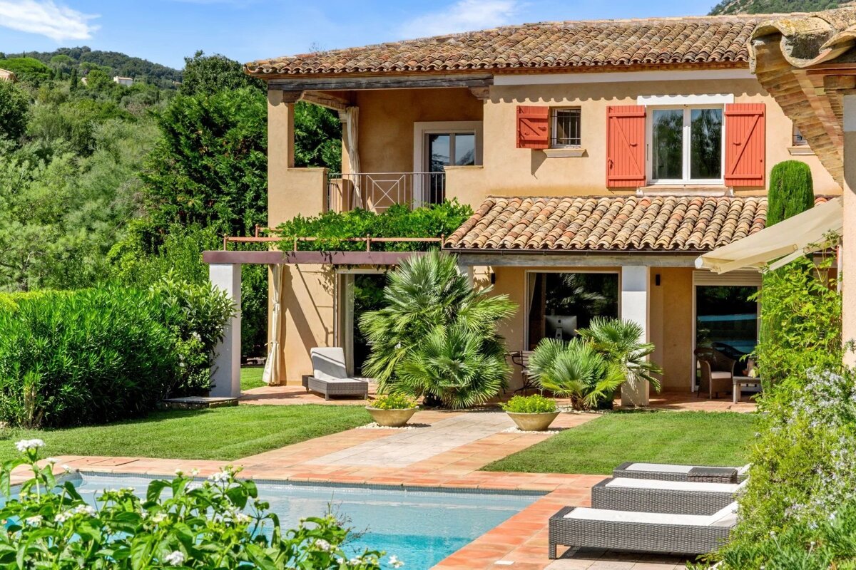 A charming Mediterranean villa with a terracotta roof, red shutters, lush garden, and a sparkling swimming pool with sun loungers.