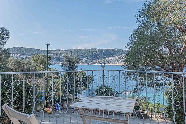Balcony with a rustic table offering a stunning view of a calm blue bay, distant hills, and coastal town under a clear sky.