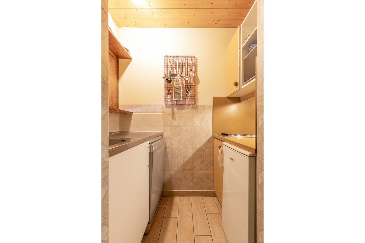 A narrow galley kitchen features a wooden ceiling, tiled walls, a sink, stove, microwave, fridge, and hanging utensils on a grid.