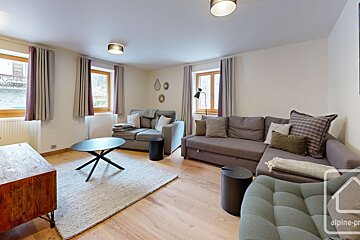 A cozy living room with two sofas, a coffee table on a rug, wooden floors, and windows with curtains, creating a warm ambiance.