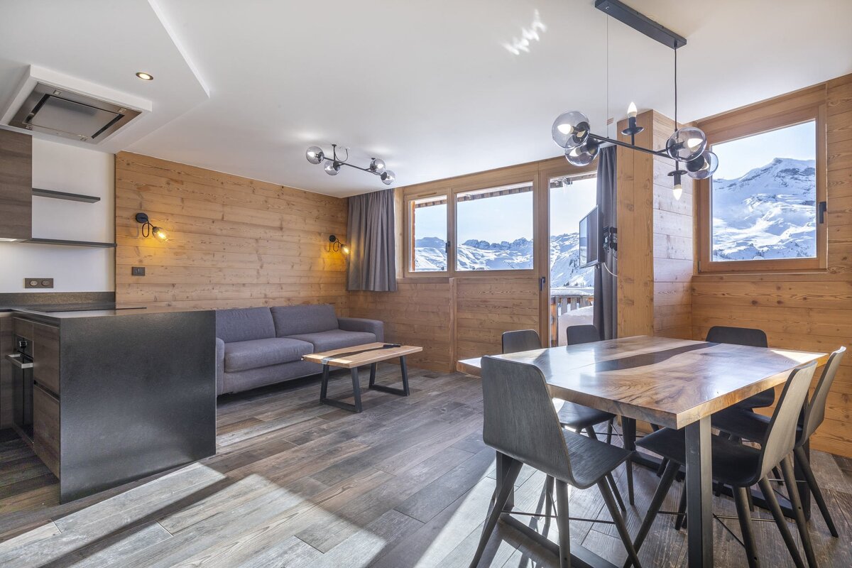 Modern ski chalet living/dining area with wood paneling, contemporary furniture, and large windows revealing stunning snowy mountain views.