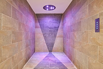 A modern, tiled shower room with warm and dark grey tiles forming a V-pattern. Purple LED lights from the ceiling and a control panel create a calming ambiance.