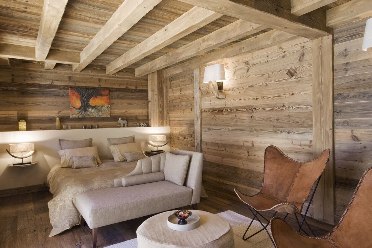 Cozy, wood-paneled chalet bedroom with exposed beams, a comfortable bed, chaise lounge, and rustic leather chairs.