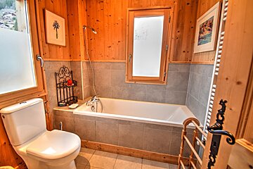 A warm, rustic bathroom featuring wooden walls, a white toilet, and a bathtub with a shower. Grey tiled lower walls, a frosted window, and decor complete the space.