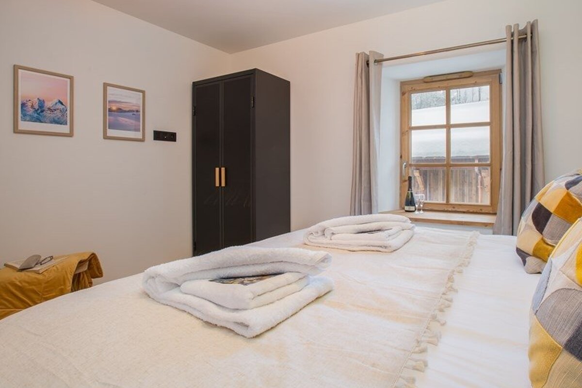 Cozy bedroom featuring a bed with folded towels, a dark wardrobe, and a wooden window looking out onto a snowy scene.