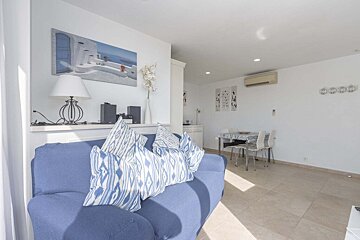 A bright, minimalist living room with a blue sofa, Santorini-style art, a dining table, and white walls, creating a clean, sunny space.