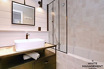 A modern bathroom features a white rectangular sink on a dark vanity, white shiplap walls, a black-framed mirror, and a tiled shower-tub.