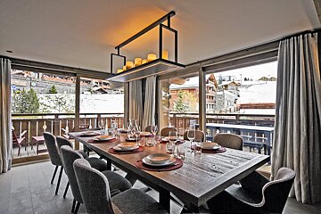 Chic dining room with a long wooden table, set for a meal. Floor-to-ceiling windows offer panoramic views of a snowy mountain village and ski chalets.