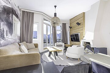 Modern alpine living room with mountain views, featuring a tan sofa, white armchairs, wooden accents, and a cozy fireplace.