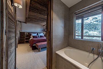 A cozy, wood-paneled bedroom is seen through a doorway, adjacent to a tiled bathroom with a tub and a window offering a snowy mountain vista.